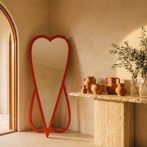 May include: A heart-shaped, red-framed full-length mirror stands against a beige wall. A stone console table displays terracotta vases and a glass vase with greenery. The room has an arched doorway.