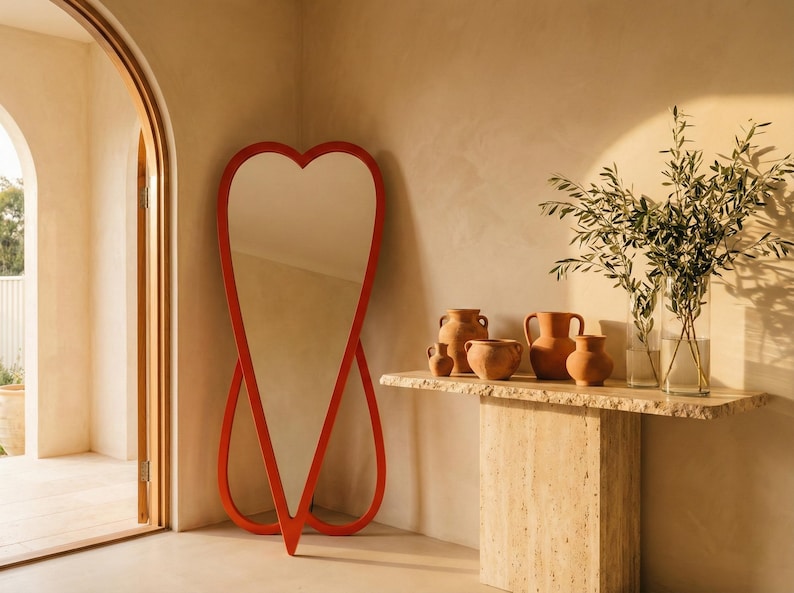 May include: A heart-shaped, red-framed full-length mirror stands against a beige wall. A stone console table displays terracotta vases and a glass vase with greenery. The room has an arched doorway.