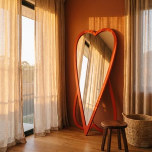 May include: A large, heart-shaped mirror with a vibrant orange frame stands against an orange wall. The mirror reflects a window with sheer curtains. A small wooden stool and a woven basket sit nearby on a wooden floor. The room is bathed in warm sunlight.