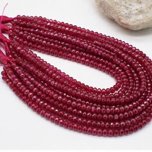Pode incluir: Um fio de contas de pedra preciosa vermelho-escuras, facetadas e redondas, dispostas em forma de U. As contas estão enfiadas e terminadas com uma borla rosa-escura a condizer numa extremidade. A superfície de cada conta reflete a luz.