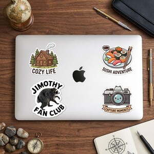 May include: Four vinyl stickers on a laptop: a log cabin with the text COZY LIFE, a sushi platter with the text SUSHI ADVENTURE, a raccoon with the text JIMOTHY FAN CLUB, and a camera with a smiling lens and the text CAPTURE MOMENTS.