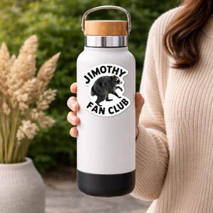 May include: A white stainless steel water bottle with a bamboo lid and a silver handle. A circular sticker on the front features a black and white raccoon illustration with the text "JIMOTHY FAN CLUB" printed in bold black capital letters.
