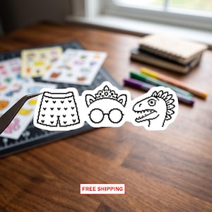 May include: A white sticker featuring three distinct pixel art designs arranged horizontally: a pair of shorts patterned with small hearts, a cat-ear tiara paired with round glasses, and the profile of a dinosaur head with a spiky crest.