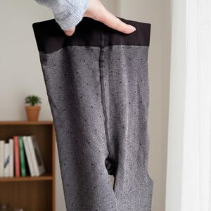 May include: Gray tights with a black waistband and small dot pattern. The tights are held up by a hand, showcasing the full length of the garment. The background includes a bookshelf and a window.