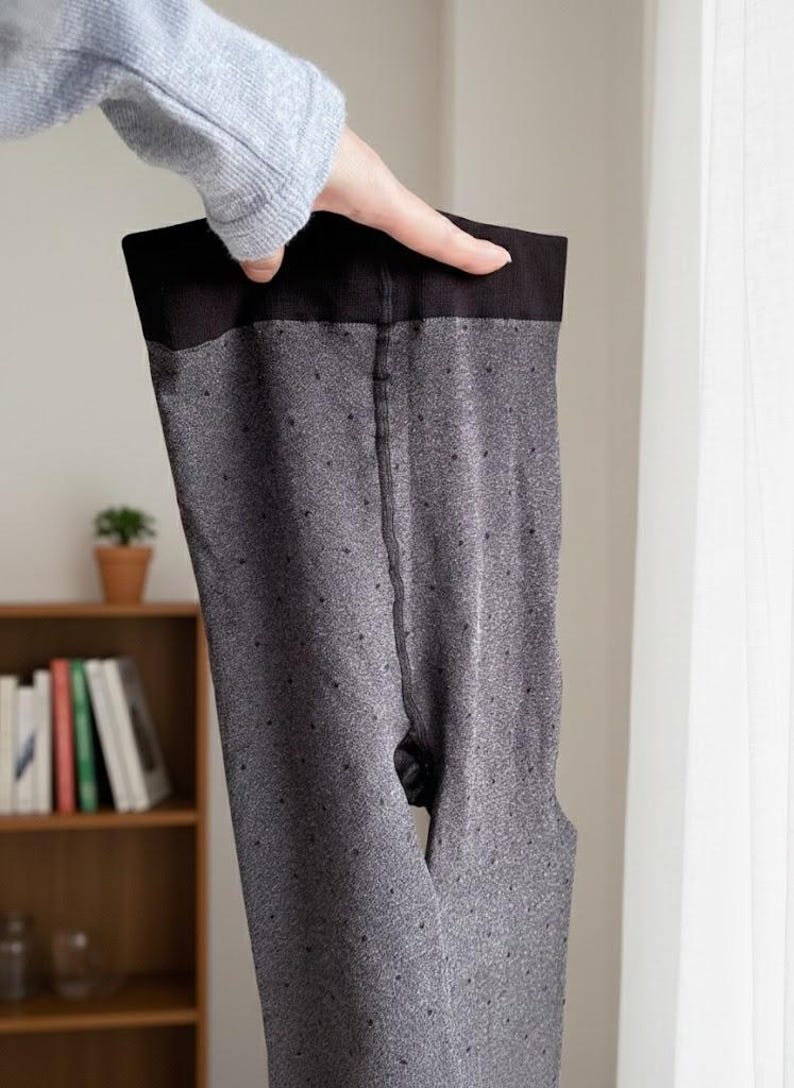May include: Gray tights with a black waistband and small dot pattern. The tights are held up by a hand, showcasing the full length of the garment. The background includes a bookshelf and a window.