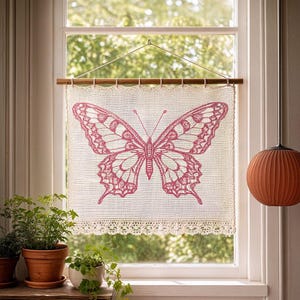 May include: A decorative window hanging featuring a large pink butterfly design on a cream-colored, open-weave fabric. The fabric is trimmed with a delicate lace border and suspended from a wooden rod.