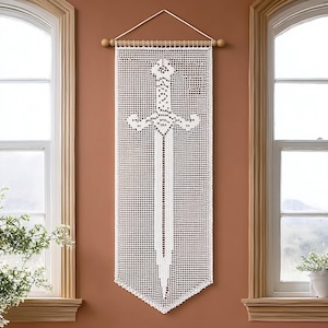 Op de afbeelding: Een witte macramé wandhanger met een zwaardontwerp. Het zwaard is het centrale kenmerk op een rechthoekig paneel, opgehangen aan een houten roede. De achtergrond heeft een rasterpatroon. De wandhanger hangt aan een bruine muur.