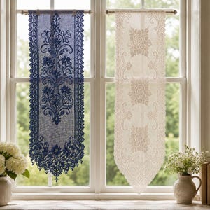 May include: Two vertical lace window panels hanging from rods. The left panel is deep navy blue with an intricate floral and scrollwork pattern. The right panel is cream-coloured with a repeating rose motif. Both feature scalloped, pointed edges.