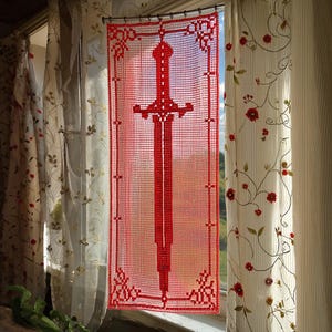 May include: A rectangular red crochet panel featuring a central sword motif with a detailed hilt and blade, enclosed within a decorative geometric border. The piece is suspended from a thin metal rod by small rings, displaying an openwork pattern.
