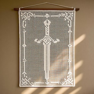 Pode incluir: Uma tapeçaria retangular de crochê branco com um motivo central de espada dentro de uma borda de grade decorativa. A peça está suspensa por uma haste de madeira escura com um cordão branco, exibindo um trabalho de agulha intrincado.