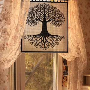 Puede incluir: Un diseño negro de árbol de la vida sobre un panel cuadrado tejido, enmarcado en negro, cuelga frente a una ventana. Las raíces y ramas del árbol están detalladas. Cortinas transparentes con estampado floral enmarcan la ventana.