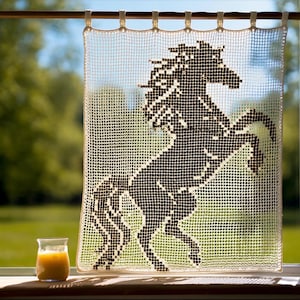 May include: A cream-colored, crocheted window curtain featuring a black silhouette of a rearing horse. The curtain hangs in front of a window with a glass of orange juice on the sill. The background is a blurred view of green trees and a blue sky.
