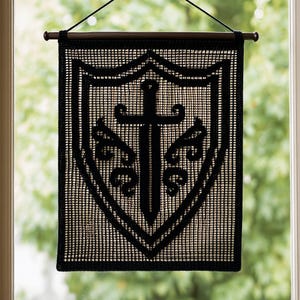 May include: Black wall hanging with a shield and sword design. The artwork is displayed on a rectangular, woven fabric. The shield and sword are outlined in black, creating a contrast against the lighter background. The banner is suspended from a wooden rod.