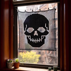 Puede incluir: Una cortina de ventana de calavera negra tejida a ganchillo cuelga en una ventana. La calavera es una característica prominente, con cuencas de los ojos, nariz y dientes detallados. La ventana está enmarcada por madera oscura, y pequeñas plantas en macetas se encuentran en el alféizar.