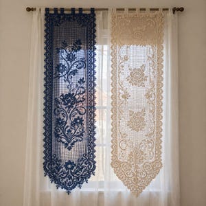 Puede incluir: Dos paneles decorativos de encaje para ventanas. Uno es azul marino con motivos florales y un borde festoneado. El otro es beige con un diseño similar. Ambos paneles cuelgan frente a una cortina blanca transparente.