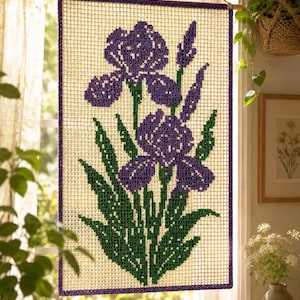 May include: A needlepoint wall hanging featuring a floral design of purple irises with green leaves and stems. The artwork is framed with a dark purple border and hangs from a wooden rod. The background is a cream-colored grid.