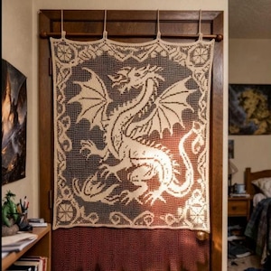 Op de afbeelding: Een crèmekleurig, gehaakt gordijn met een gedetailleerd drakenontwerp. De draak staat centraal op het gordijn, met zijn vleugels gespreid. Het gordijn hangt aan een houten roede en voegt een vleugje fantasie-decor toe aan een deuropening.