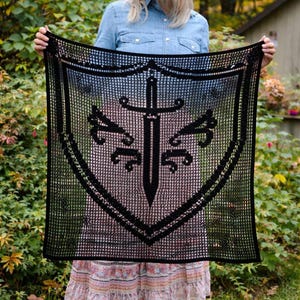 May include: A black crocheted shawl featuring a shield design with a sword in the center. The shawl is held up, showcasing the intricate openwork pattern. The design includes decorative elements and a border.