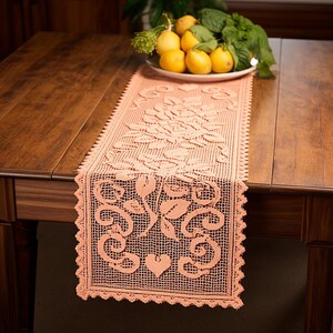 May include: A rectangular peach-colored crochet table runner featuring an intricate openwork pattern of roses, leaves, and heart shapes. The runner has a scalloped edge and is laid out flat on a dark wooden table surface with a bowl of lemons and basil.