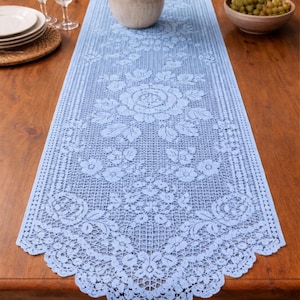 May include: A pale blue lace table runner with a floral pattern, displayed on a wooden table. The runner has a detailed design of roses and other flowers. The table is set with plates, glasses, and a vase of white flowers.