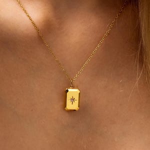 Könnte beinhalten: Goldfarbene Halskette mit rechteckigem Anhänger. Der Anhänger zeigt ein Sternmuster und hängt an einer zarten Kette. Die Halskette wird vor einem weichen, neutralen Hintergrund gezeigt.