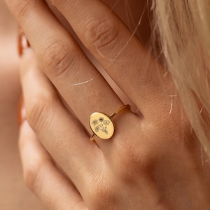 Puede incluir: Un anillo de oro con una cara ovalada adornada con un diseño floral. El anillo se lleva en un dedo y el diseño está grabado en negro. La banda del anillo es fina y sencilla.