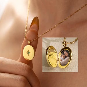 May include: A gold oval locket necklace featuring a starburst engraving with a small green stone at the center. The locket opens to reveal a photograph of a person holding a child and the engraved text Best Mom on the interior left side.