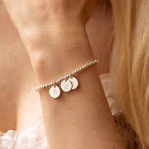 Puede incluir: Una pulsera de cuentas plateada con tres pequeños colgantes redondos. Los colgantes tienen símbolos y letras grabados. La pulsera se lleva en la muñeca y el fondo está borroso.