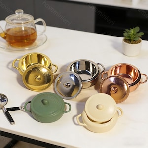 Può includere: Set di pentole in miniatura con pentole dorate, argentate, ramate, verdi e color crema con coperchi. Sono visibili anche una teiera di vetro con tè e una piccola pianta grassa. Il set è disposto su una superficie bianca.