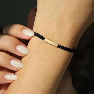 Könnte beinhalten: Ein schwarzes Kordelarmband mit einem rechteckigen, goldfarbenen Metallsteg, der mittig am Handgelenk sitzt. Der Steg ist mit dem Wort MOVE graviert und enthält einen kleinen, klaren, runden Stein zwischen den Buchstaben O und V.