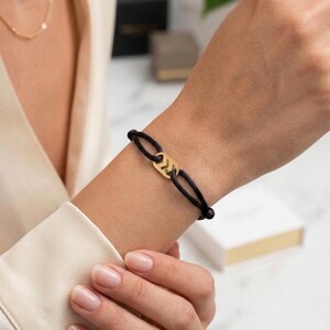May include: A black cord bracelet with a gold-colored chevron design. The bracelet is worn on a wrist, showcasing the jewelry's minimalist style. The gold accent adds a touch of elegance to the simple design.