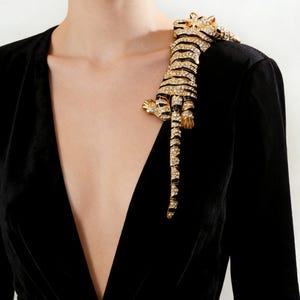 Op de afbeelding: Een goudkleurige tijgerbroche met zwarte strepen en sprankelende kristallen accenten. De broche is bevestigd aan een zwart fluwelen kledingstuk, wat een opvallend contrast creëert. De tijger is afgebeeld in een klimmende pose.