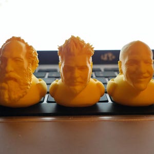 May include: Three yellow rubber duck-shaped figurines, each featuring a distinct sculpted human face. The left figure has a full beard, the center figure has short spiky hair and a goatee, and the right figure has a smooth head and a mustache.