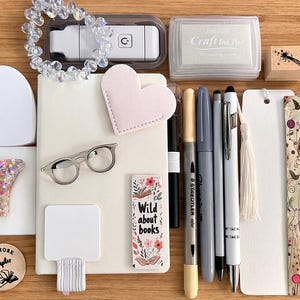 May include: A collection of stationery and accessories, including a notebook, pens, a bookmark, and a heart-shaped notepad. The image also features a small pair of glasses, a stamp, and a decorative bracelet. The items are arranged on a wooden surface.