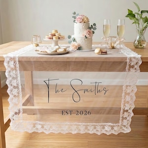 Puede incluir: Un camino de mesa transparente con ribete de encaje blanco y el texto "The Smiths EST 2026" se muestra sobre una mesa de madera. La mesa está decorada con una tarta de boda, macarons, copas de champán y otros elementos decorativos.