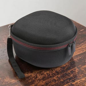 May include: A black, rounded carrying case with a textured surface. The case has a red zipper and a black wrist strap. The case is designed to protect headphones or other small electronics.