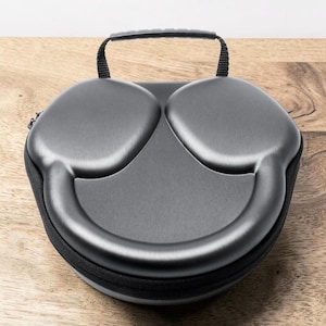 Könnte beinhalten: Ein graues Kopfhörer-Etui mit schwarzem Reißverschluss und kleinem Griff. Das Etui ist für Over-Ear-Kopfhörer konzipiert und hat eine glatte, geschwungene Oberfläche. Das Etui liegt auf einer Holzoberfläche.