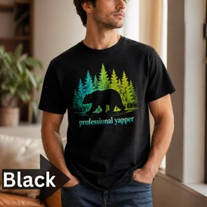 May include: Black t-shirt with a bear silhouette walking in front of a forest with blue and yellow-green trees. The text "professional yapper" is below the bear. The shirt is made of a soft material.