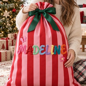 May include: A large fabric gift sack featuring vertical red and pink stripes, cinched at the top with a dark green satin ribbon bow. The name MADELINE is spelled out in colourful, textured, crocheted letters, accented by a small crocheted butterfly and flower.