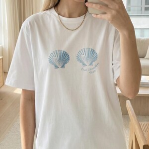 May include: White t-shirt with two light blue seashell designs. The text "East Hampton 2024" is printed below the seashells. The shirt has short sleeves and a crew neck.