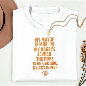 Op de afbeelding: Een wit t-shirt met ronde hals en oranje tekst: MY MAYOR IS MUSLIM. MY BAGEL'S JEWISH. THE POPE IS ON OUR SIDE, KNICKS IN FIVE. Onder de tekst staat een klein oranje en blauw basketbal-logo met het woord KNICKS.