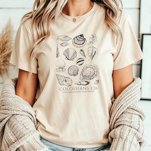 May include: A beige short-sleeved t-shirt featuring a black line-art graphic of various seashells arranged in a cluster. Below the shells, the text reads COLOSSIANS 1:16 FOR BY HIM ALL THINGS WERE CREATED in a simple, clean, black sans-serif font.