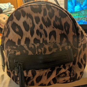 May include: A small backpack featuring a brown and black leopard print pattern. The bag includes a top handle, a main zippered compartment, and a smaller front zippered pocket with a black leather-like trim and a long black zipper pull.