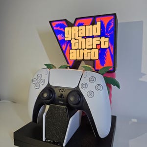 May include: A white and black video game controller resting on a white stand, positioned in front of a backlit sign featuring the Grand Theft Auto V logo with a palm tree graphic in orange, purple, and black colors, all mounted on a black base.