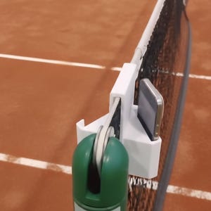 May include: A tennis net with a white support system and a green and white device attached. A smartphone is mounted on the device. A yellow tennis ball is in the background on a brown court.