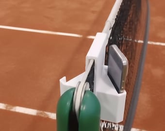 Op de afbeelding: Een tennisnet met een wit ondersteuningssysteem en een groen en wit apparaat bevestigd. Een smartphone is op het apparaat gemonteerd. Een gele tennisbal is op de achtergrond op een bruine baan.