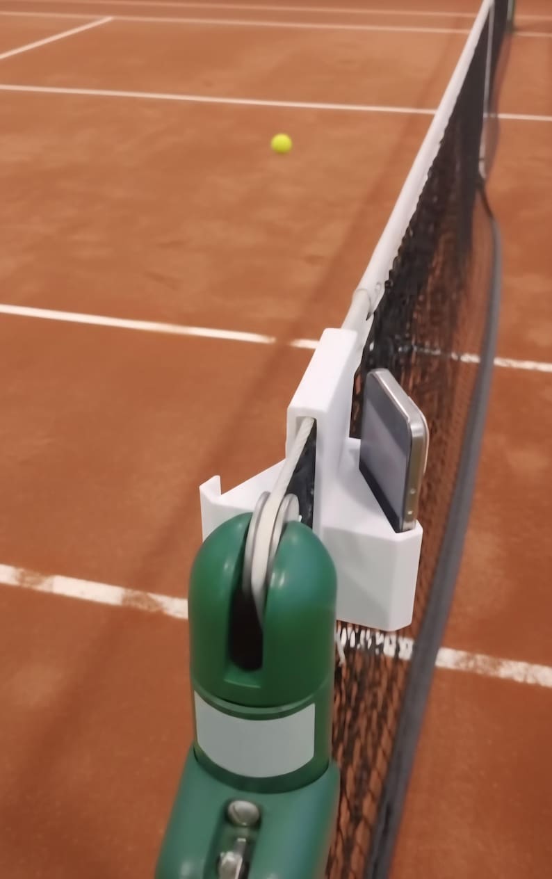 May include: A tennis net with a white support system and a green and white device attached. A smartphone is mounted on the device. A yellow tennis ball is in the background on a brown court.