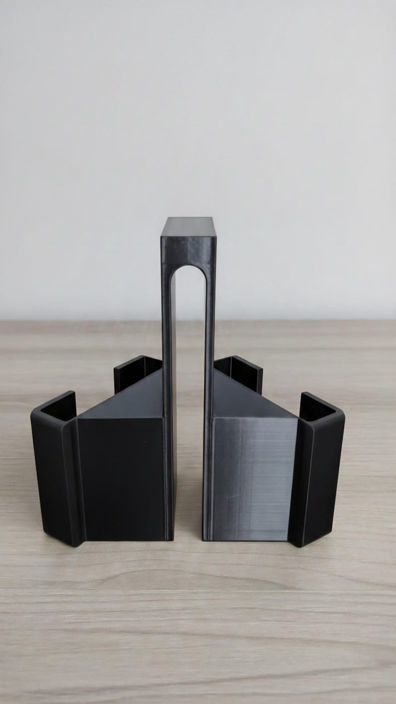 May include: Black geometric bookends. The bookends have a modern design with a rectangular base and a tall, arched support. The bookends are made of a solid, matte black material and are displayed on a light wood surface.