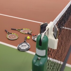 May include: A tennis court scene with tennis rackets, a yellow tennis ball, and a net. A white phone holder is attached to the net, holding two phones. The court has a green and brown surface.