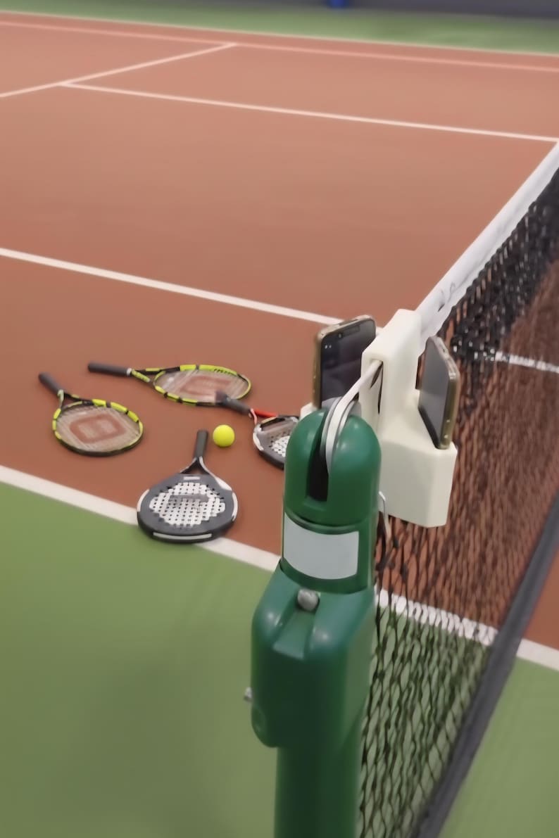 May include: A tennis court scene with tennis rackets, a yellow tennis ball, and a net. A white phone holder is attached to the net, holding two phones. The court has a green and brown surface.